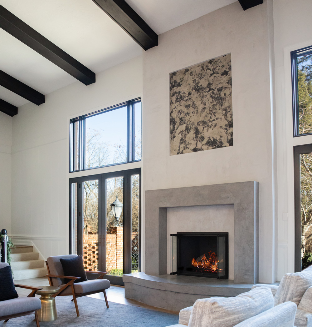 Modern living room with a minimalist concrete fireplace, large windows, and exposed ceiling beams.